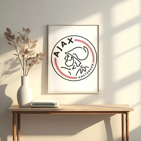 Logo del Ajax enmarcado, con sombra de luz natural, sobre una mesa con una jarra y un libro.