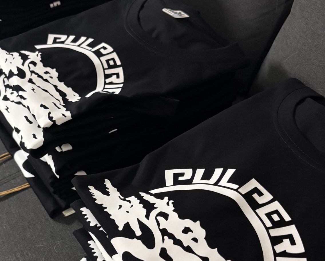 Camisetas negras apiladas con el logo "PULPERIA" en blanco.